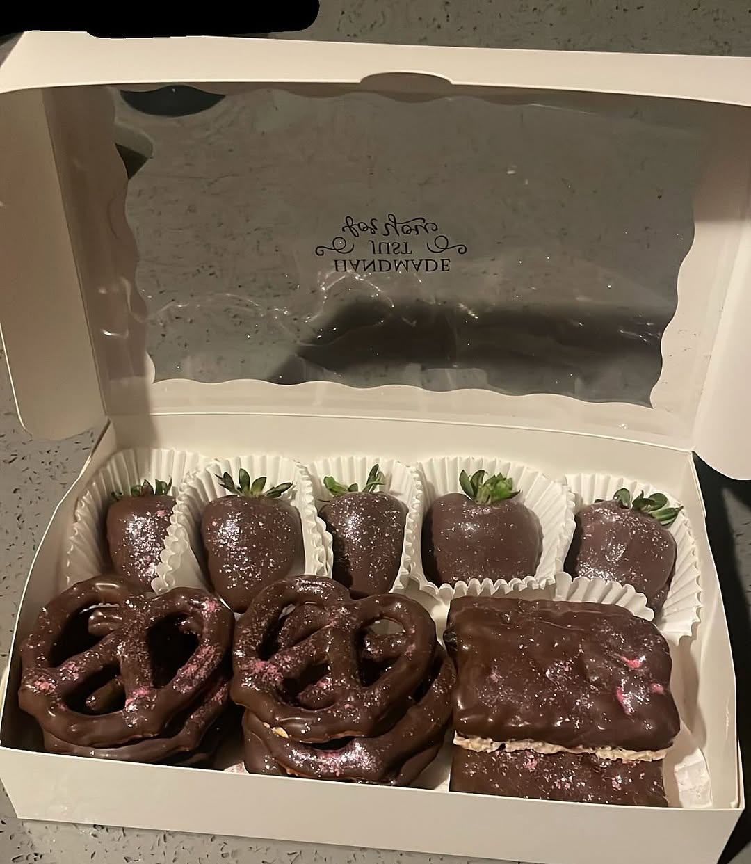 Assorted dark chocolate treats with pink rose shimmer dust, including four strawberries, two pretzels, and a Rice Krispie bar in a white bakery box.