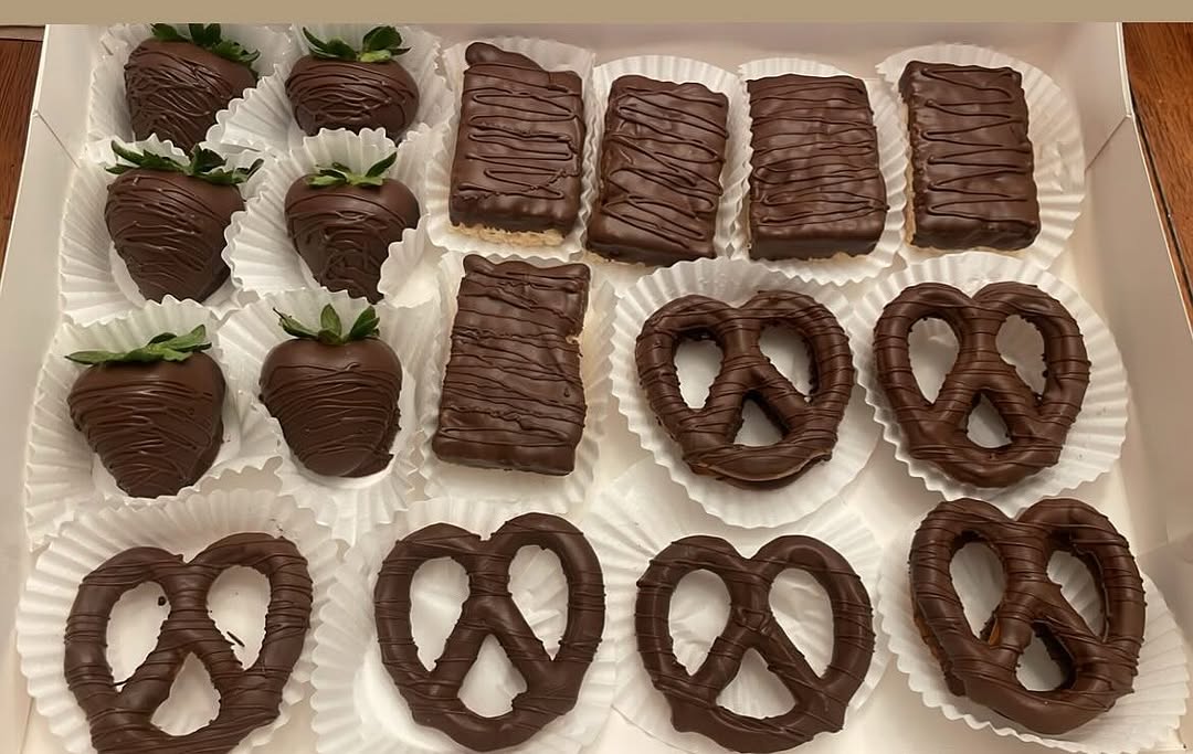 Large party platter box of assorted chocolate-covered strawberries, Rice Krispie bars, and pretzels with decorative chocolate drizzle patterns.