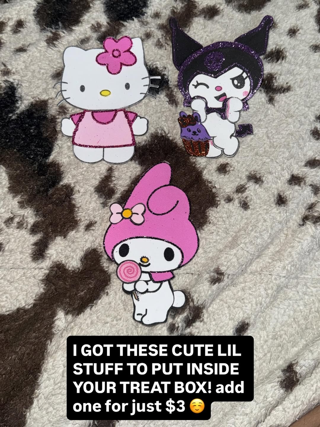 Three glittery Sanrio character cutout decorations of Hello Kitty, Kuromi, and My Melody displayed on cow-print fabric, available as treat box add-ons.