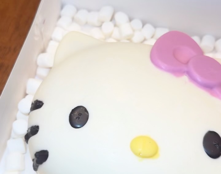 Close-up of a white chocolate Hello Kitty breakable shell showing the smooth coating, pink dimensional bow, black eyes, yellow nose, and piped whiskers.