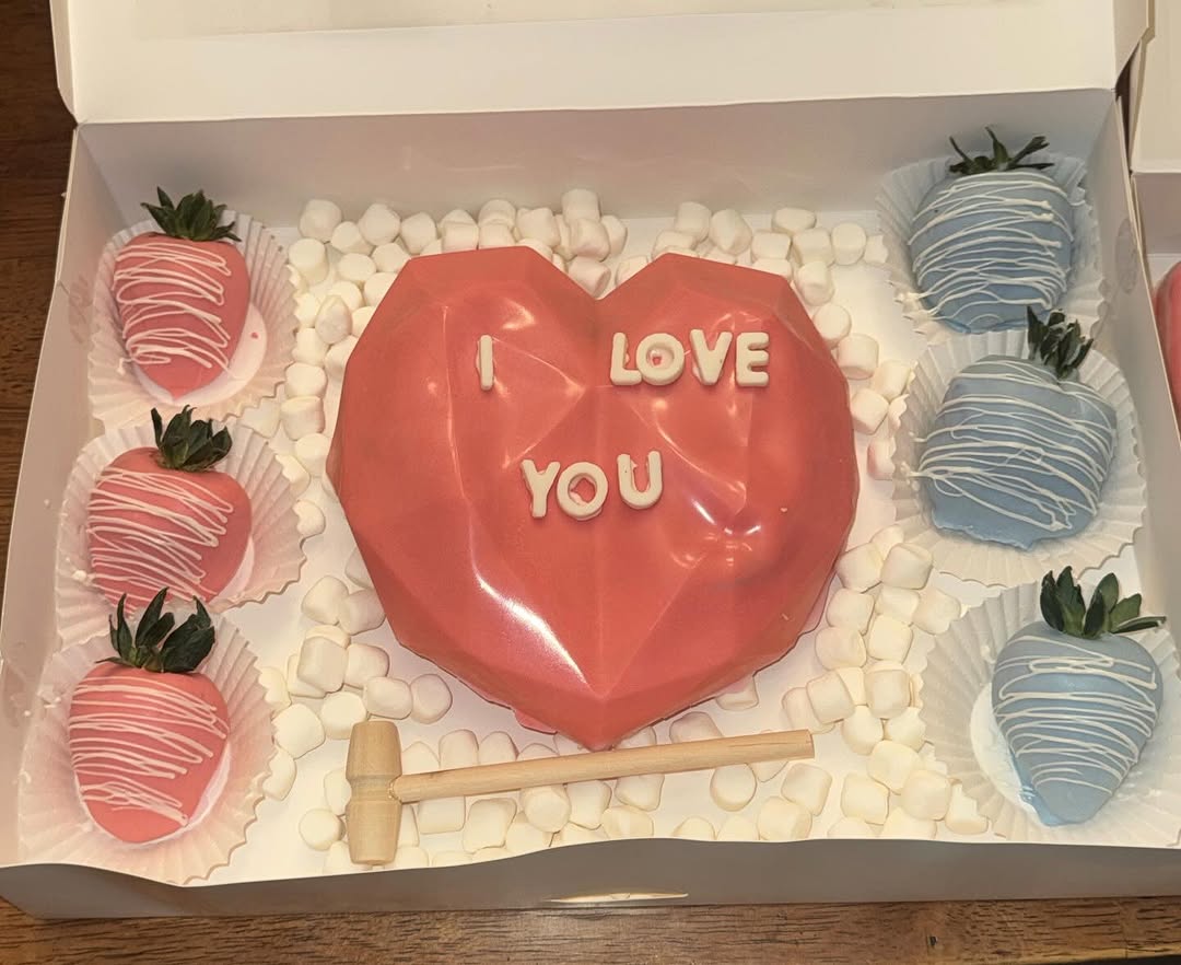 Valentine's breakable pink heart with I LOVE YOU text, flanked by three pink and three baby blue drizzled strawberries and a wooden mallet.