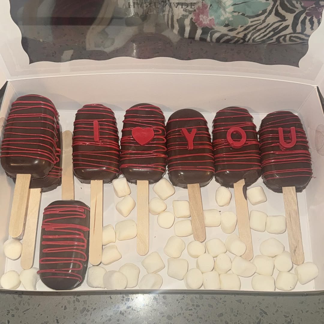 Seven dark chocolate Valentine's cakesicles with red drizzle and three spelling I HEART YOU in red fondant letters, standing in marshmallows.