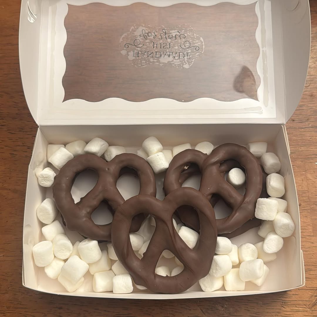Three large dark chocolate-covered pretzels in a white bakery box cushioned with mini marshmallows.