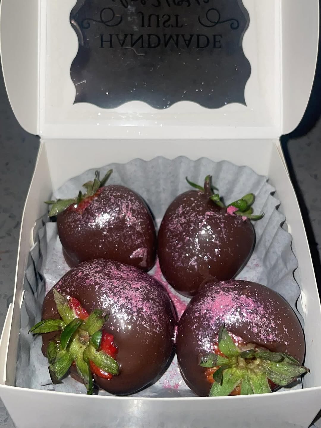 Four dark chocolate-covered strawberries generously dusted with vibrant pink magenta shimmer powder in a small white bakery box.
