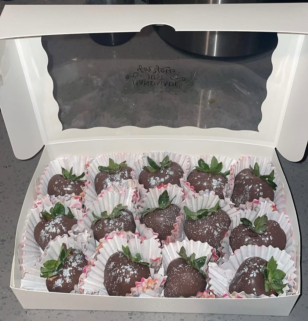 A dozen dark chocolate-covered strawberries with subtle white shimmer in decorative pink floral cupcake liners inside a white bakery box.