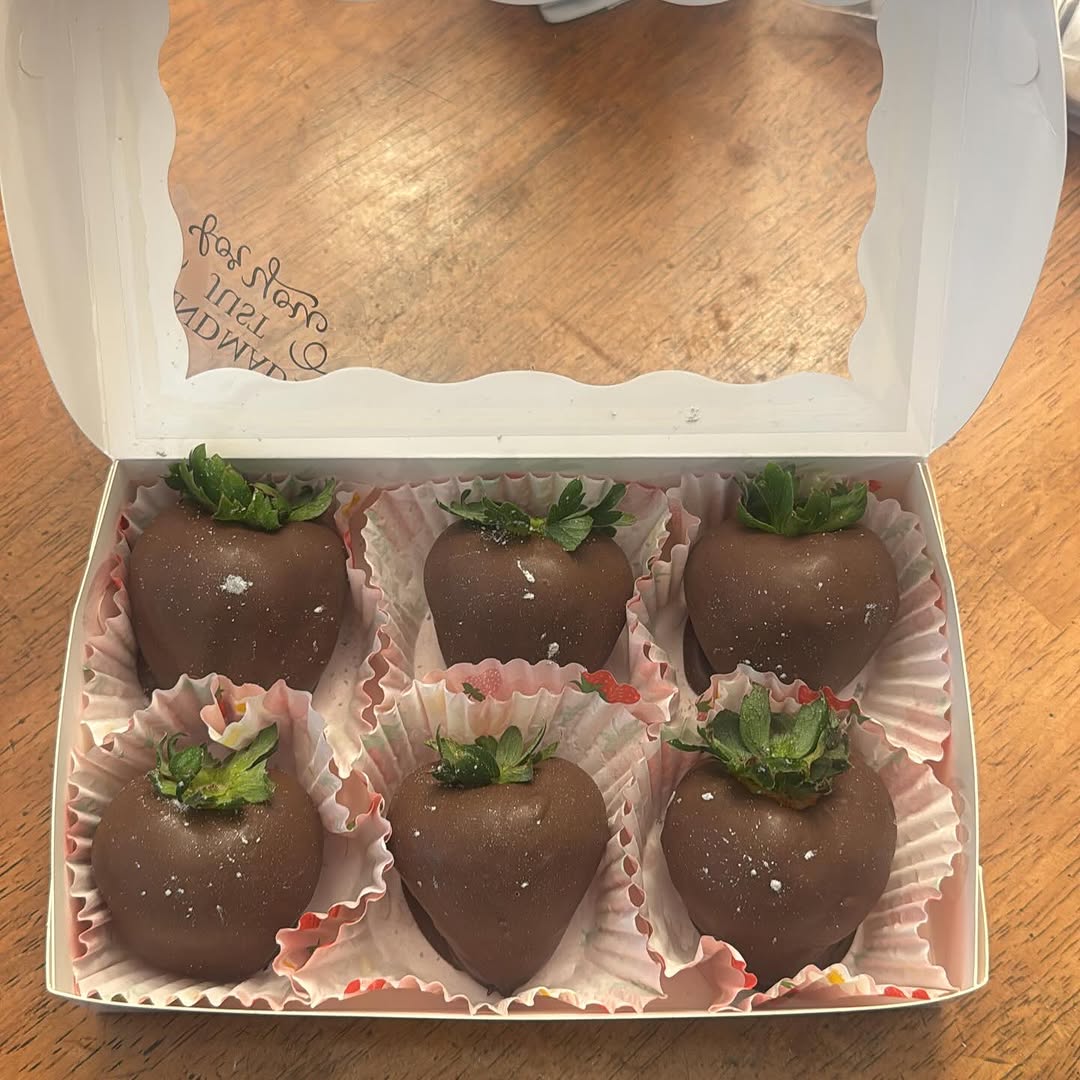 Half-dozen dark chocolate-covered strawberries with gold iridescent shimmer dust in pastel pink paper liners, an elegant gift presentation.