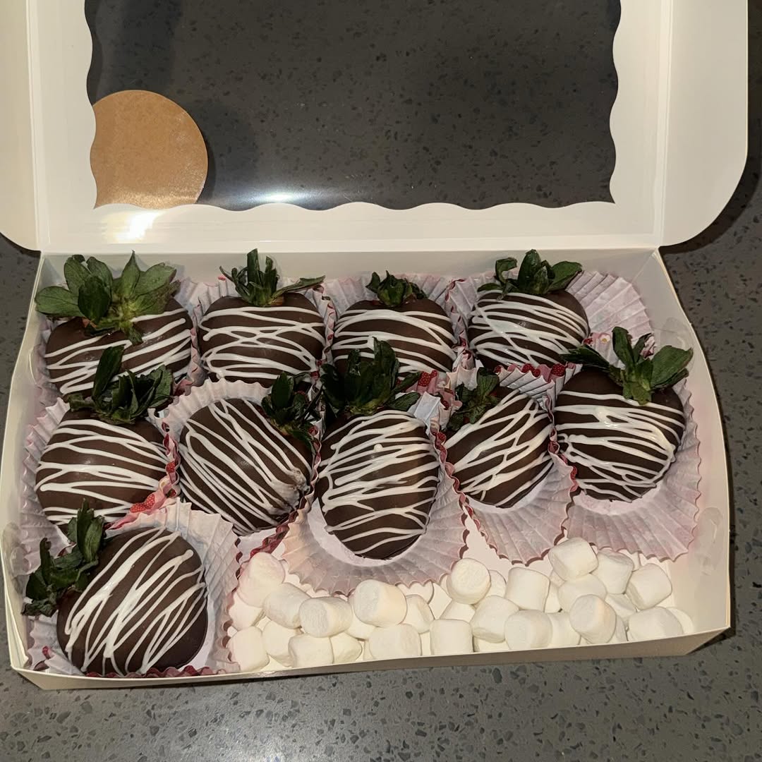 A dozen dark chocolate strawberries with white chocolate drizzle stripes in pink-tinted liners, accented with mini marshmallows in the corner.