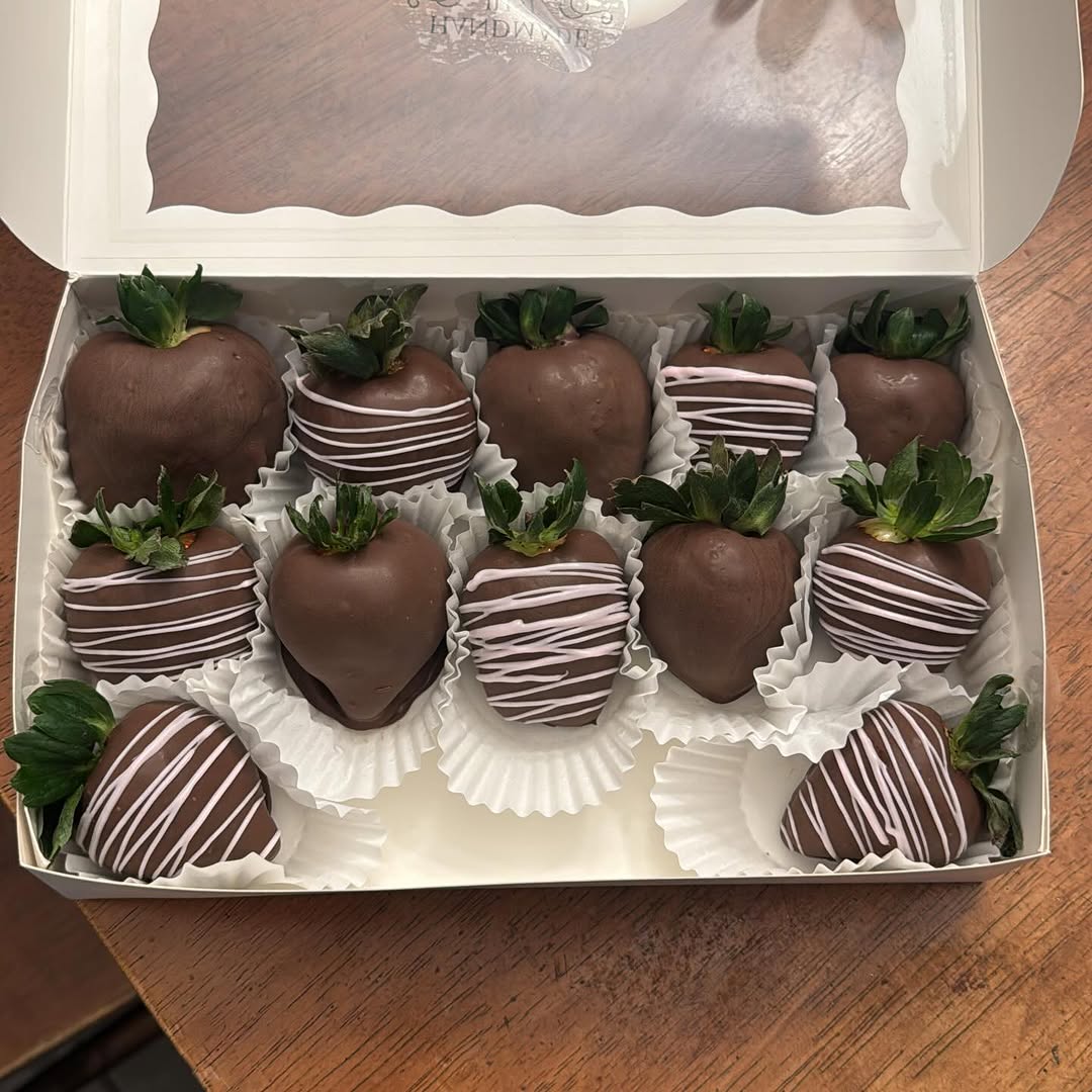 Eleven dark chocolate-covered strawberries with delicate white and pink diagonal drizzle lines in a full white bakery box.